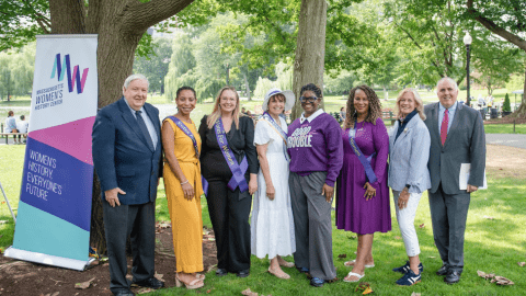 8 speakers from Women's Equality Day event standing on the Boston Common next to an MWHC banner