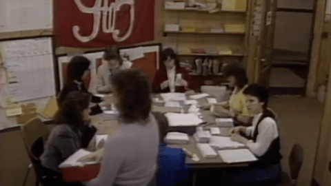 group of women around a table stuffing envelopes. A red flag on the wall says "9to5" in white text.