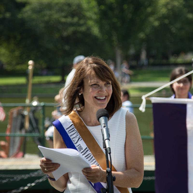 Woman wearing sash speaks outside at microphone.