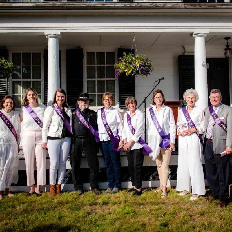 Large group of adults stands outside wearing purple sashes.