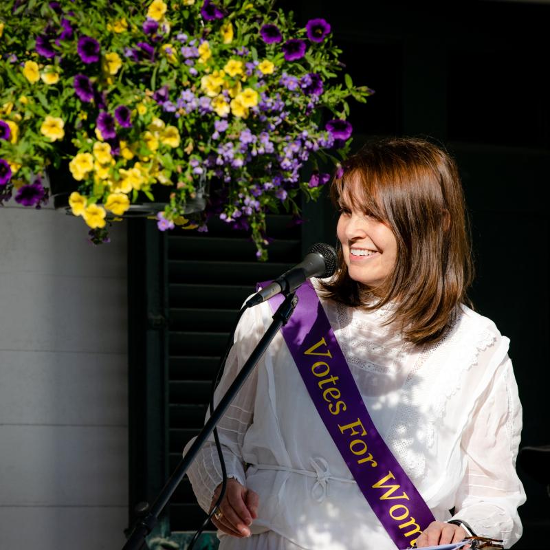 Woman stands outdoors wearing purple sash.