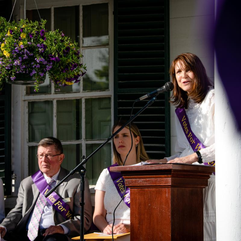 Woman wearing purple sash stands outside speaking at lectern.