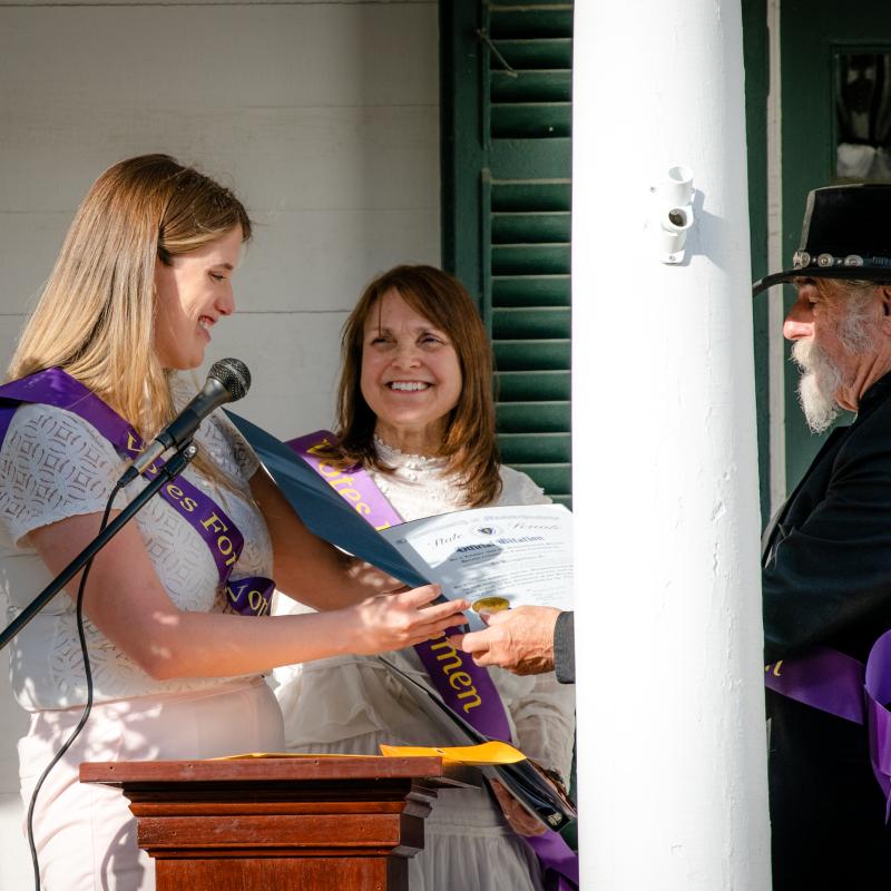 Woman wearing purple sash stands outside speaking at lectern.