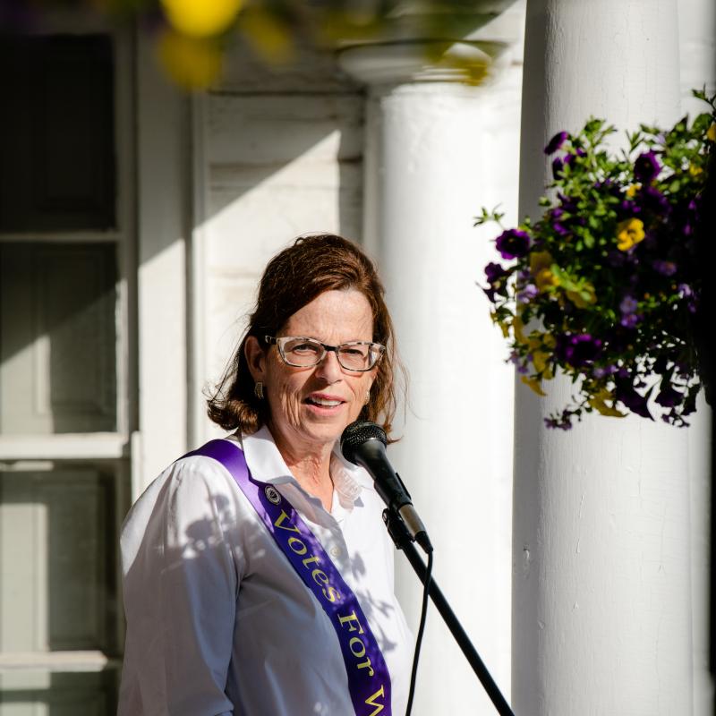 Woman wearing purple sash stands outside speaking at lectern.