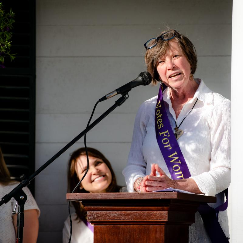 Woman wearing purple sash stands outside speaking at lectern.