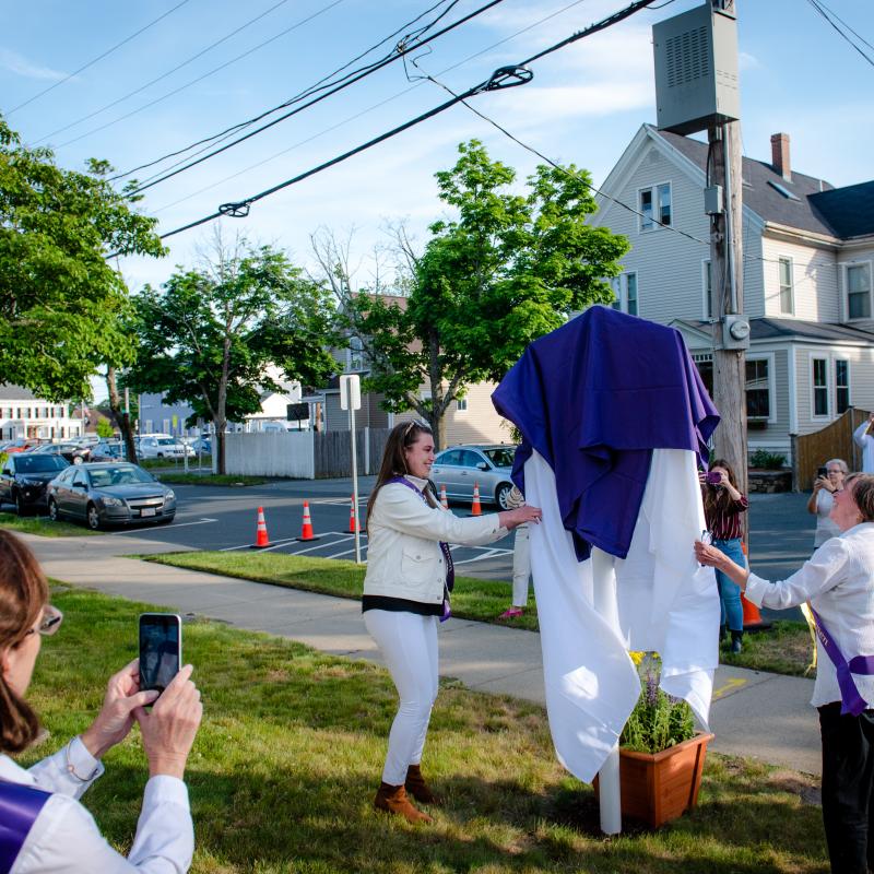 Woman stands next to sign covered in purple cloth.