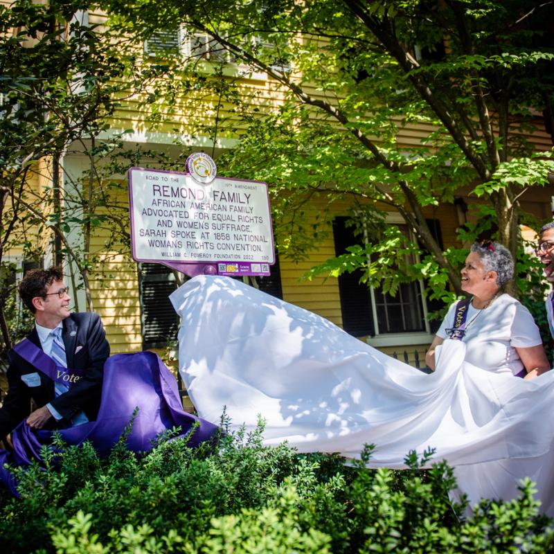 Three people pull cover off white and purple sign.