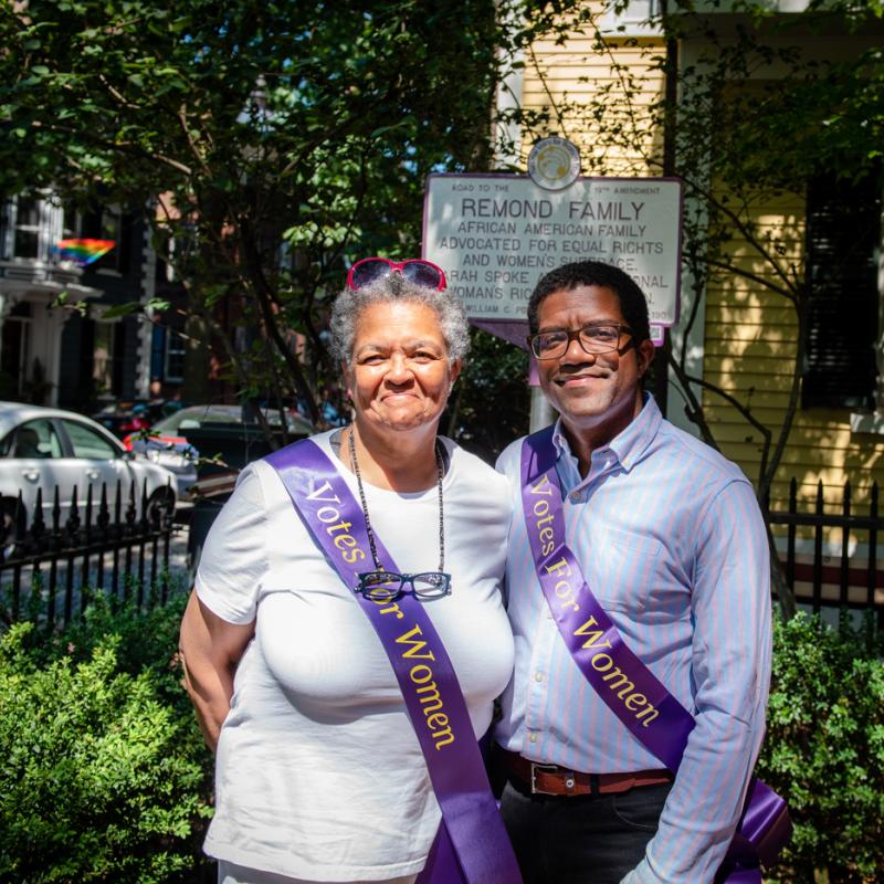 Two adults stand in front of purple and white sign.