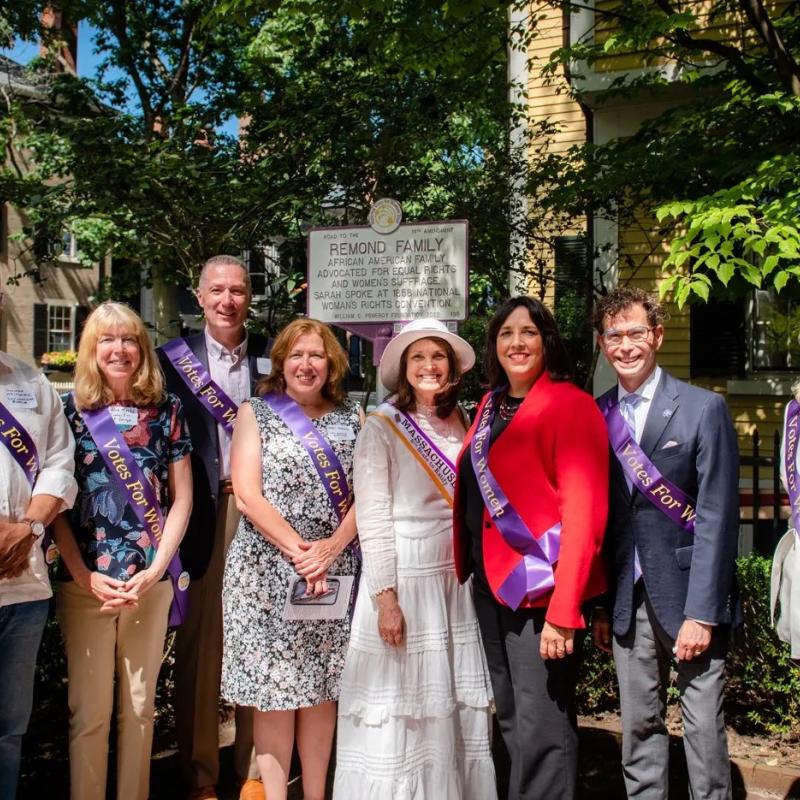 Nine people stand together wearing purple sashes and smiling.