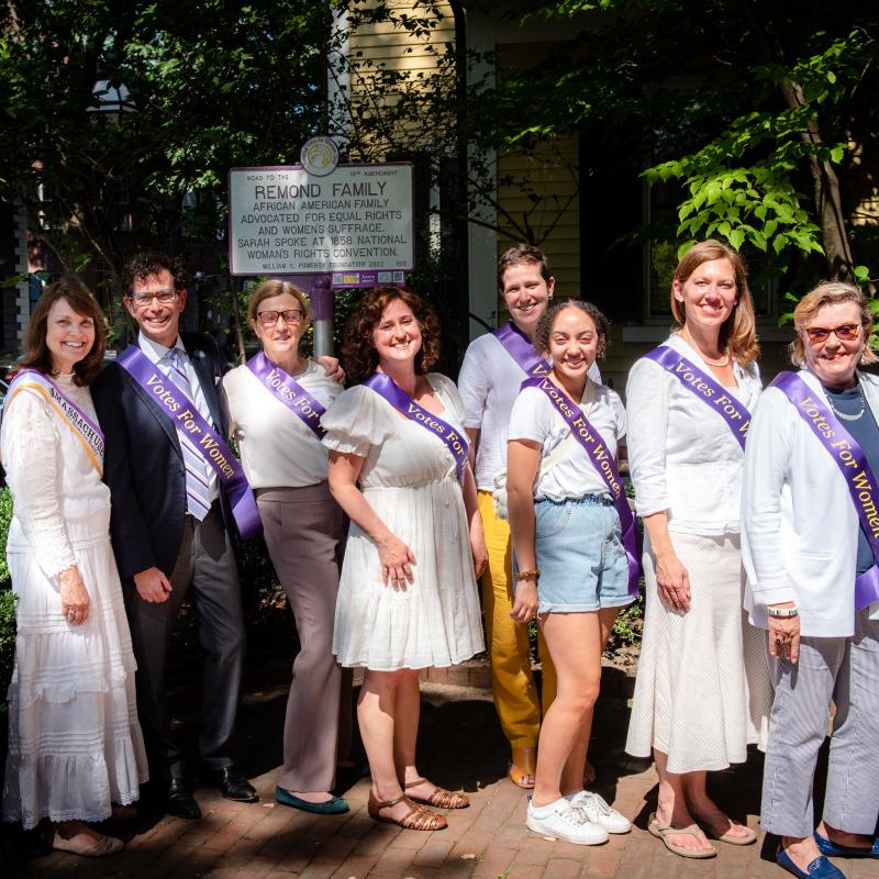 Eight people stand in front of purple and white sign.