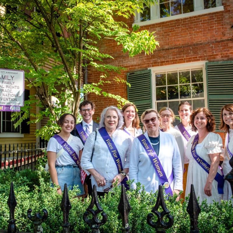 Group of people stand near purple and white sign.