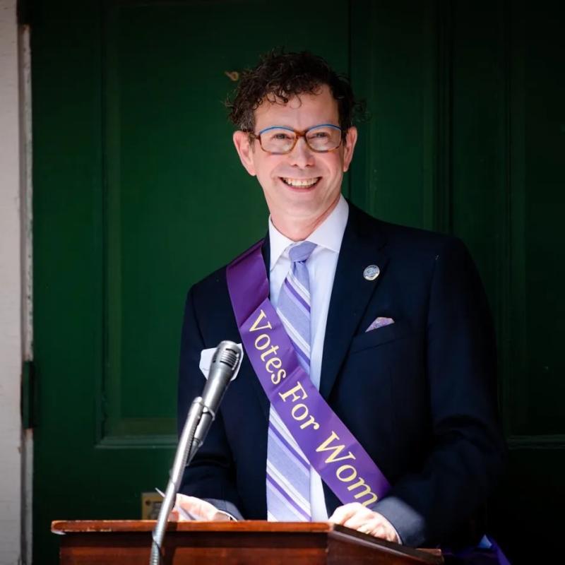 Man wearing purple sash stands outside speaking at lectern.