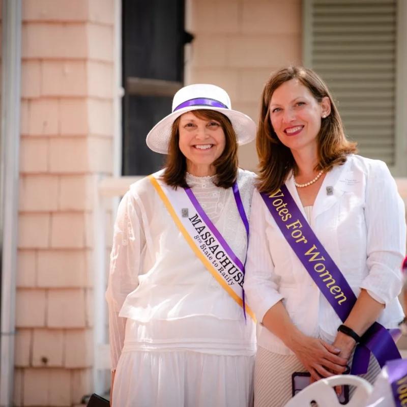 Two women wearing purple sashes stand outside smiling.