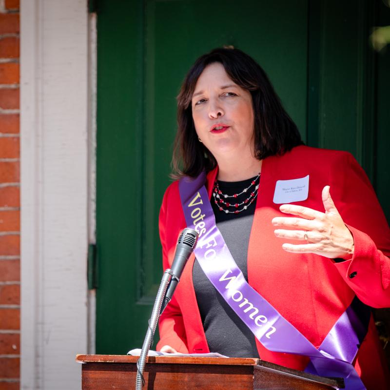 Woman wearing purple sash speaking at podium.