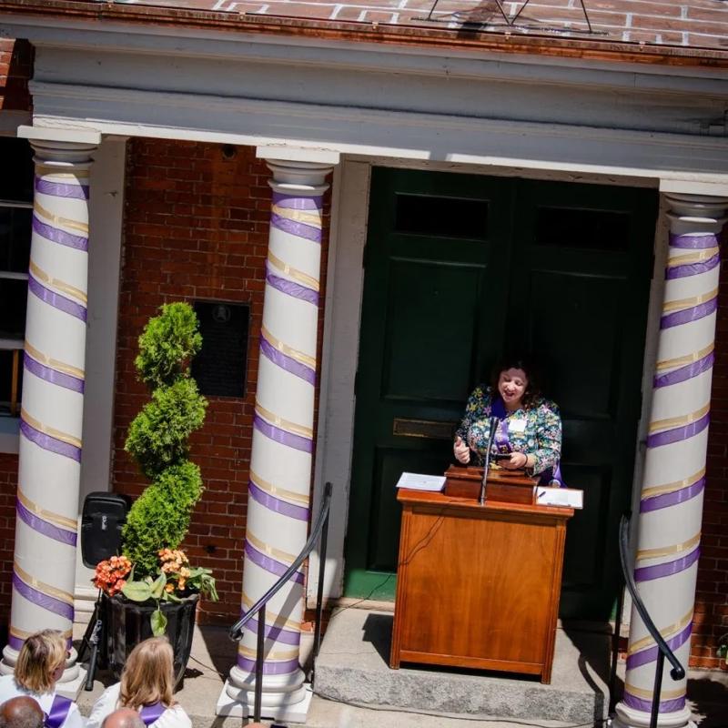 Woman wearing purple sash stands at podium.