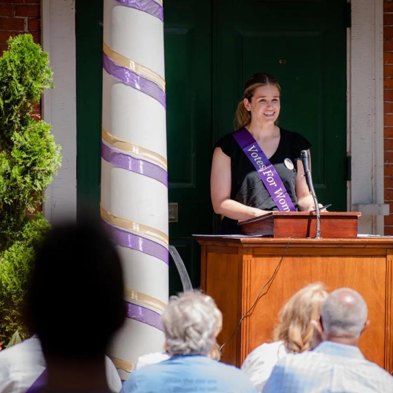 Woman wearing purple sash stands outside speaking at lectern.