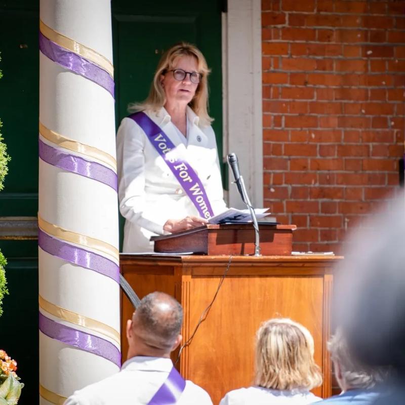 Woman wearing purple sash stands outside speaking at lectern.