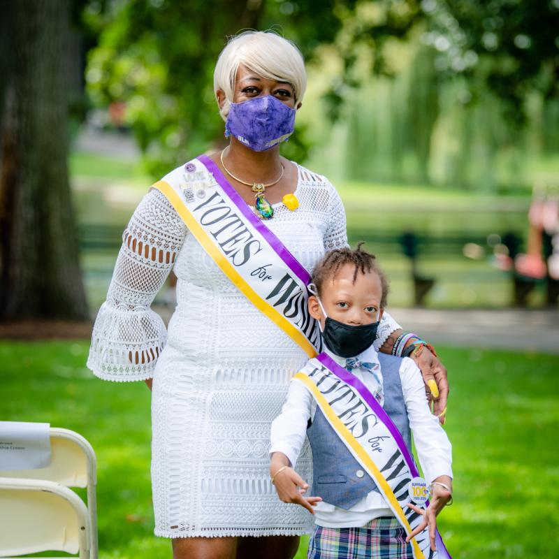 Woman and girl stand outside wearing masks and purple sashes.