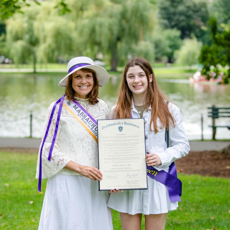 Two women stand outside wearing purple sashes.