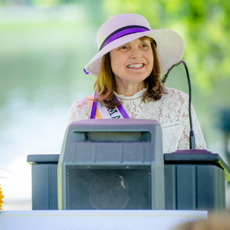 Woman stands at podium outside wearing purple sash.