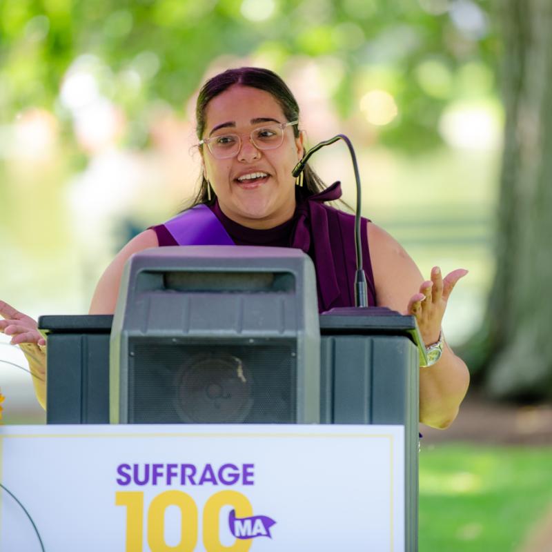 Woman wearing purple sash stands outside speaking at lectern.