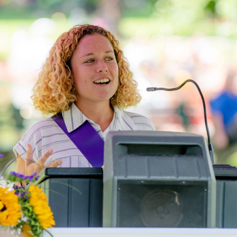 Woman wearing purple sash stands outside speaking at lectern.