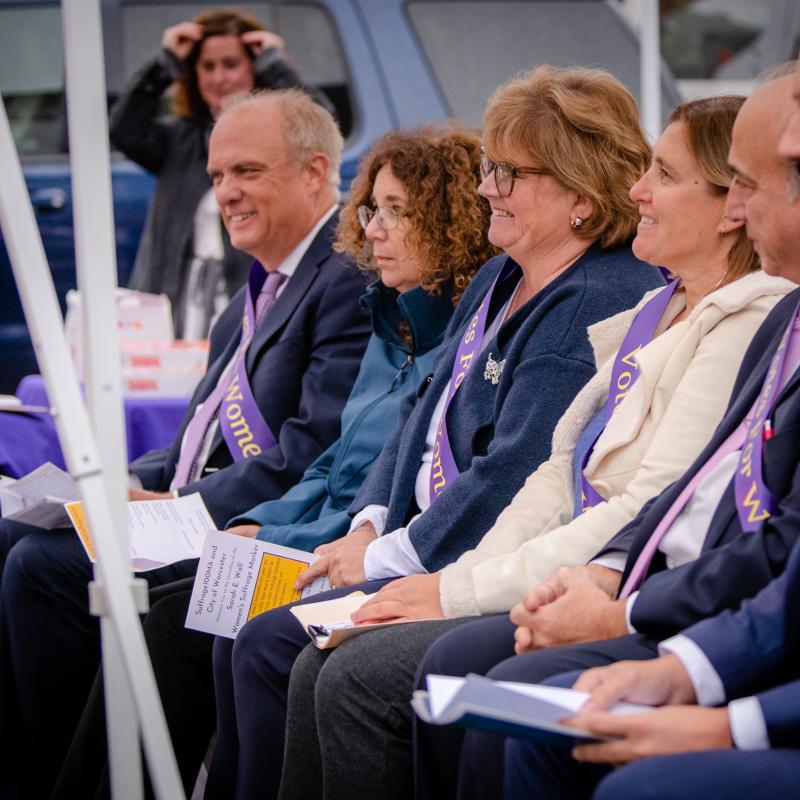 Group of people wearing purple sashes sitting in chairs.