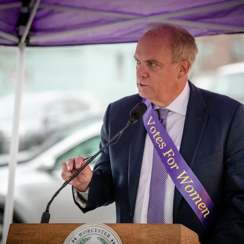 Man wearing purple sash stands outside speaking at lectern.