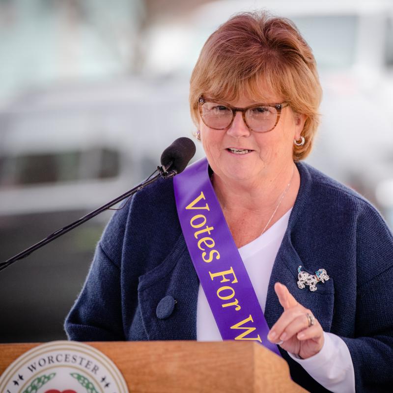 Woman wearing purple sash speaking at podium.