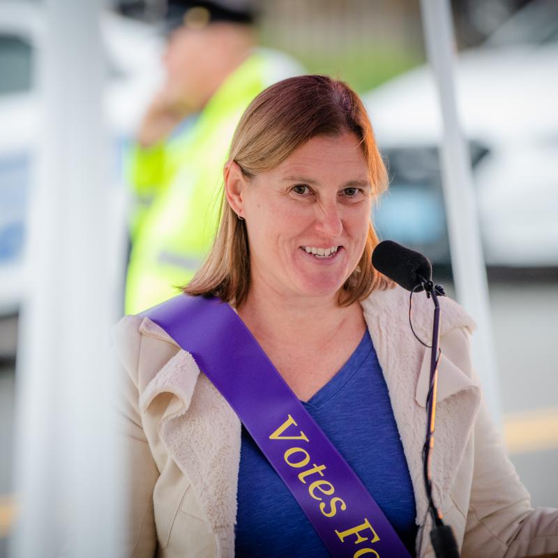 Woman wearing purple sash stands outside speaking at lectern.