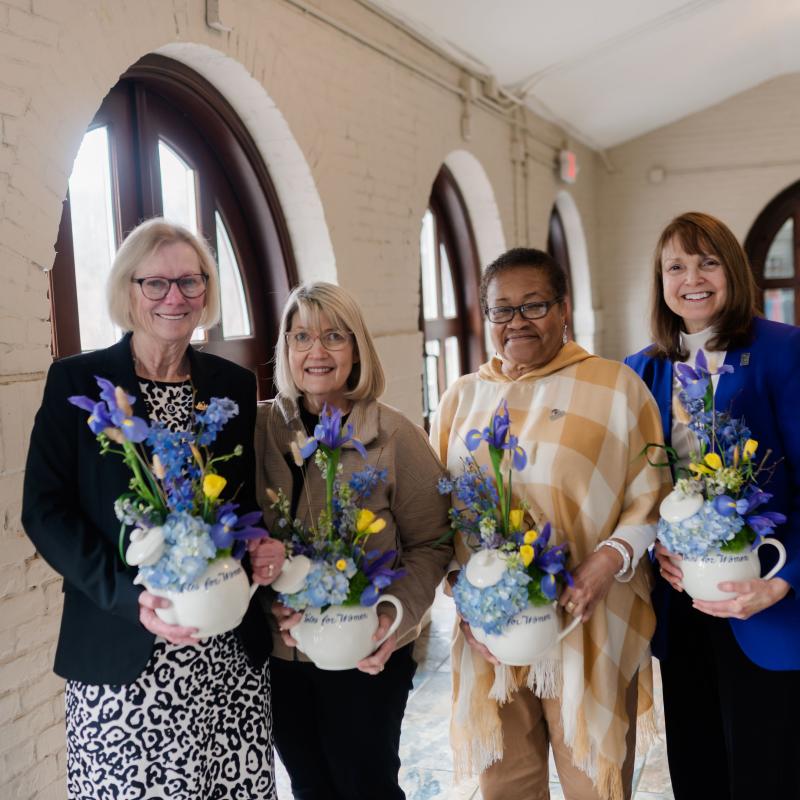 Four women stand indoors smiling and holding flowers.