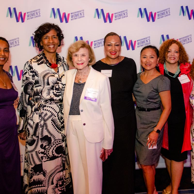 Seven women stand in front of Massachusetts Women's History Center's step and repeat.