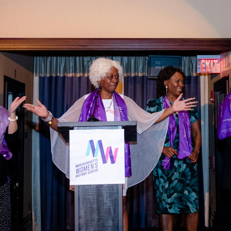 Three women wearing purple scarves stand onstage.
