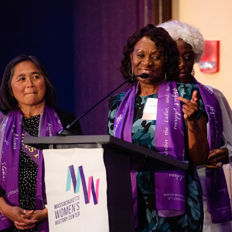 Three women wearing purple scarves stand onstage.