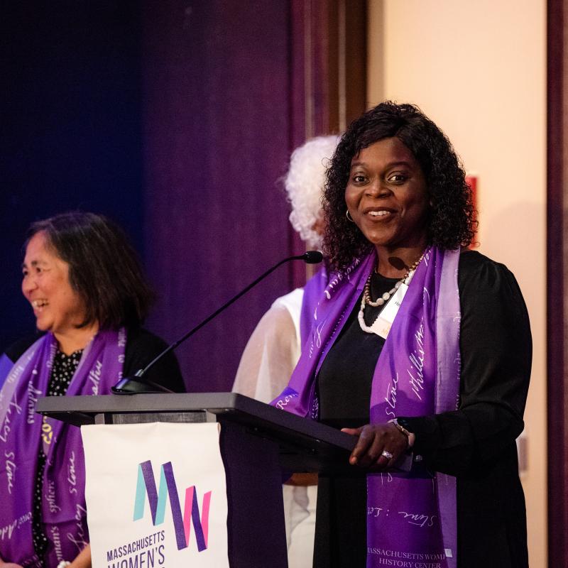 Three women wearing purple scarves stand onstage.