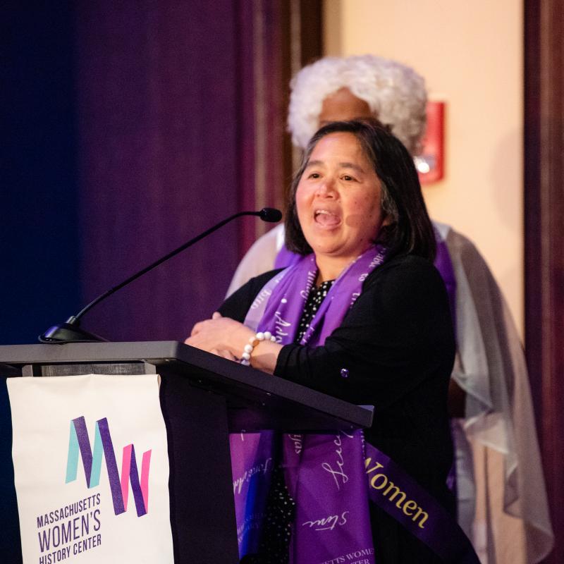 Woman in purple scarf stands onstage speaking.