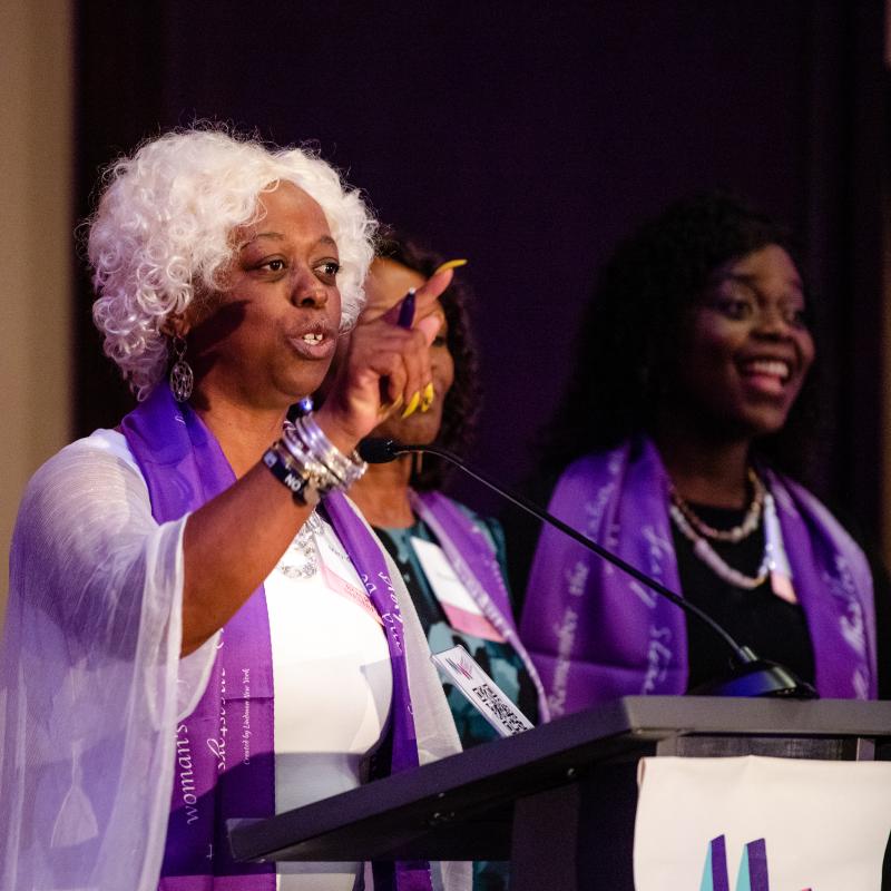 Three women wearing purple scarves stand onstage.