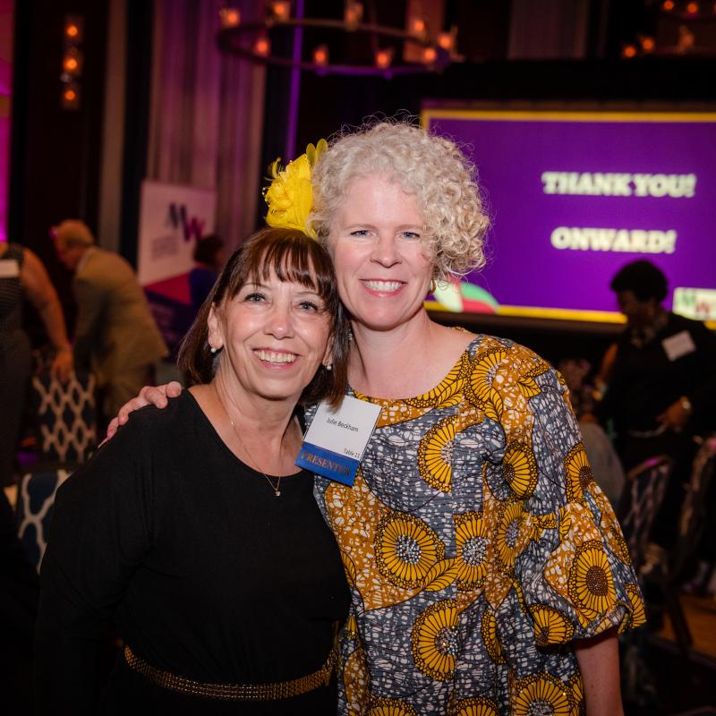 Two women stand smiling indoors.