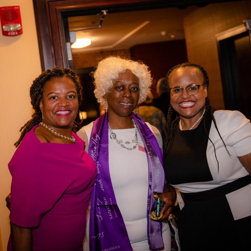 Three women stand indoors smiling.