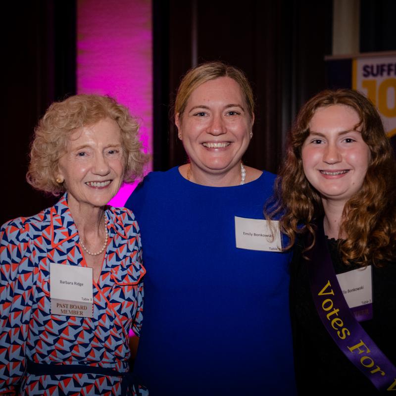 Three women stand indoors smiling.