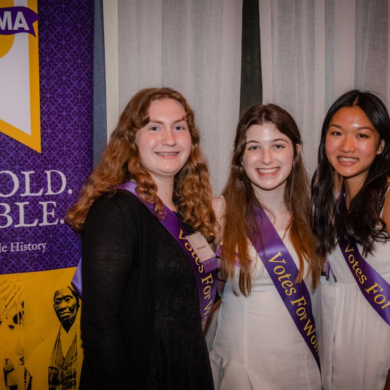 Three women in purple sashes stand indoors smiling.