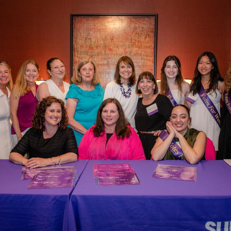 10 women stand and sit by purple table.