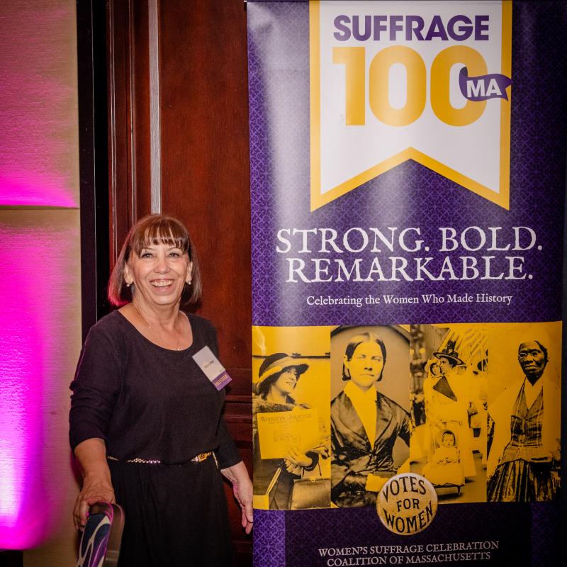 Woman stands in front of Suffrage100MA banner.