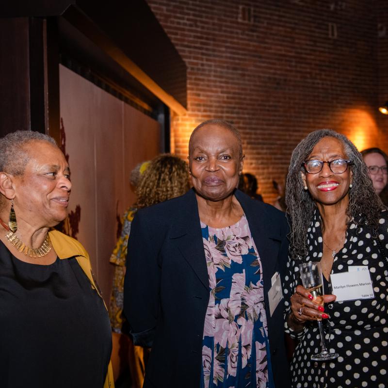 Three women stand indoors smiling.