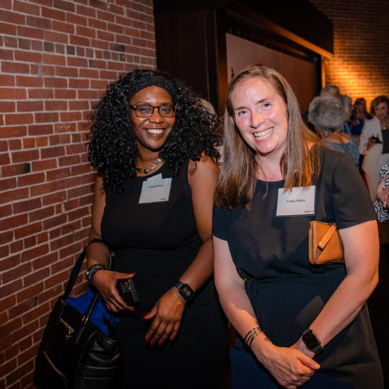 Two women stand indoors smiling.