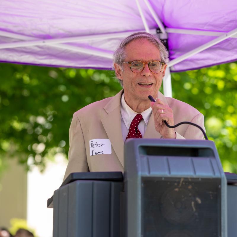 Man wearing purple sash stands outside speaking at lectern.