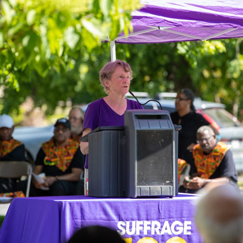 Woman wearing purple sash speaking at podium.