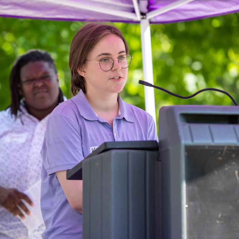 Woman wearing purple sash stands outside speaking at lectern.