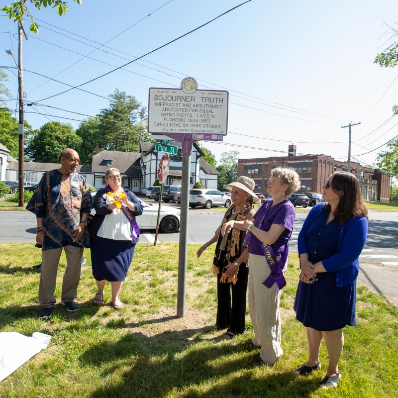 Group of people wearing purple sashes stand outside smiling next to Sojourner Truth marker.