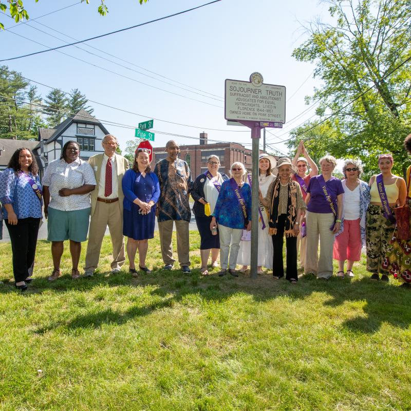 Group stands smiling outside next to purple and white sign.